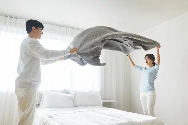 침실에서 구스이불을 털어 복원력을 유지하는 부부 – 구스다운 관리 요령